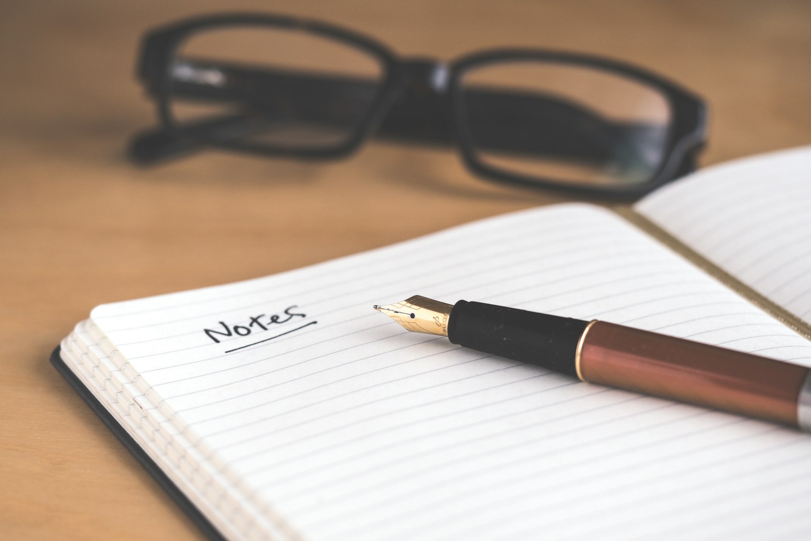 Open notebook with the word 'Notes' written in fountain pen, copper-coloured pen and reading glasses on a wooden desk — practical learning.